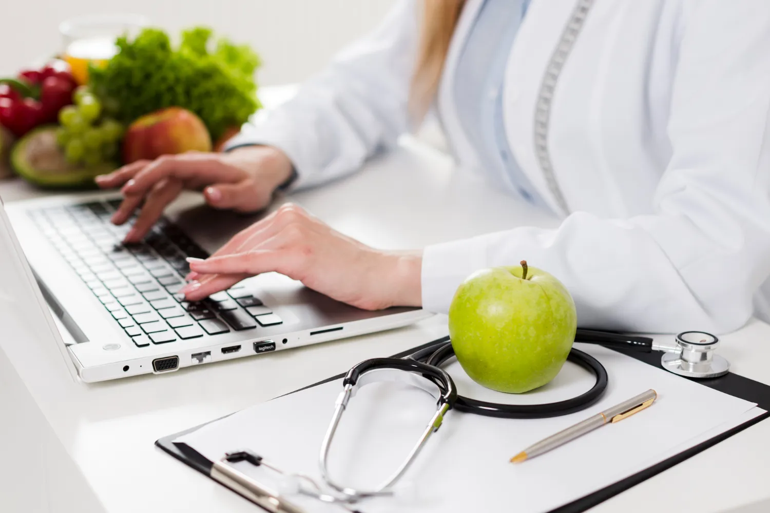 Médico escribiendo en un ordenador portátil, con una manzana verde y un estetoscopio en la mesa, rodeado de alimentos saludables como frutas y verduras.
